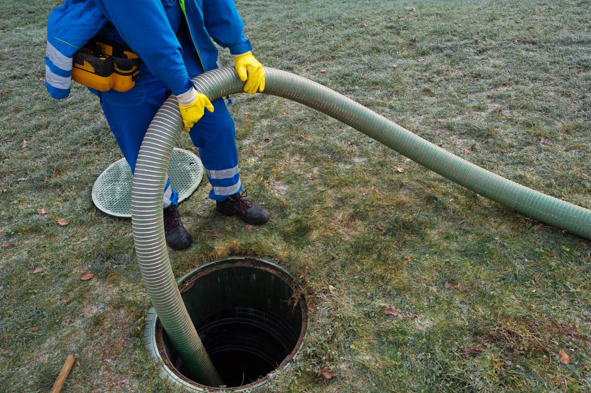 A septic service worker lowers a large, thick, corrugated pumping hose into an open round septic tank.